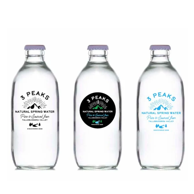 Three bottles of 3 Peaks natural spring water, each with different colored labels: black, white, and blue, featuring mountain and sun graphics.
