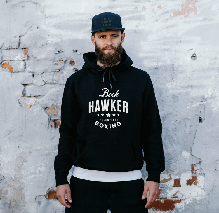 A man with a beard wearing a black hoodie with 'Beck Hawker Boxing' logo and a black cap standing against a weathered gray brick wall.
