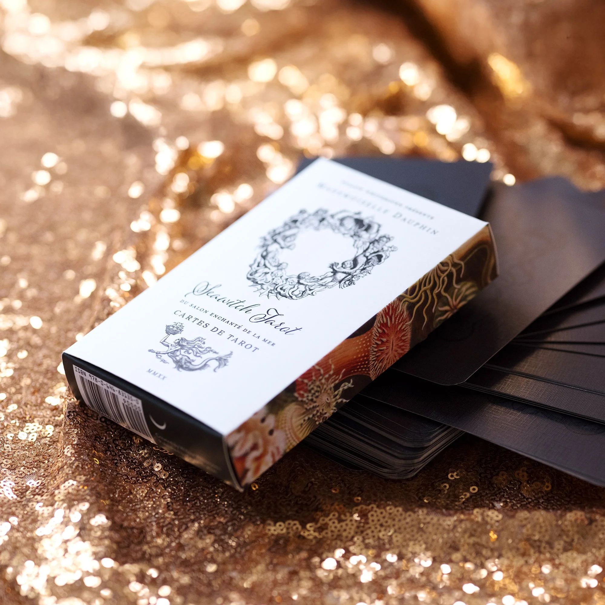 A deck of tarot cards named 'Seawitch Deck' resting on a gold sequin fabric. The box features a black and white illustration of a wreath and a stylized creature, with dark black edges and decorative artwork on the sides.
