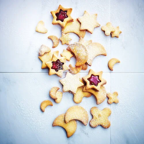 Assorted cookies in various shapes, some with jam filling, arranged on a white surface with powdered sugar sprinkled around.