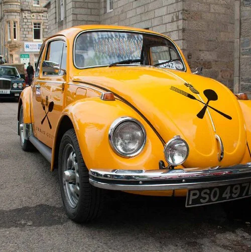Yellow vintage car with black crossing spoon and fork symbols on the doors and hood parked on a city street.