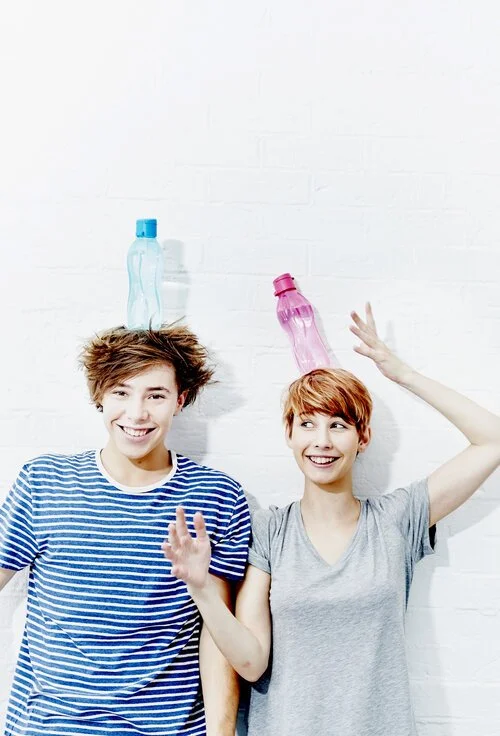 Two young women with short hair balancing empty plastic bottles on their heads and smiling against a white brick wall.