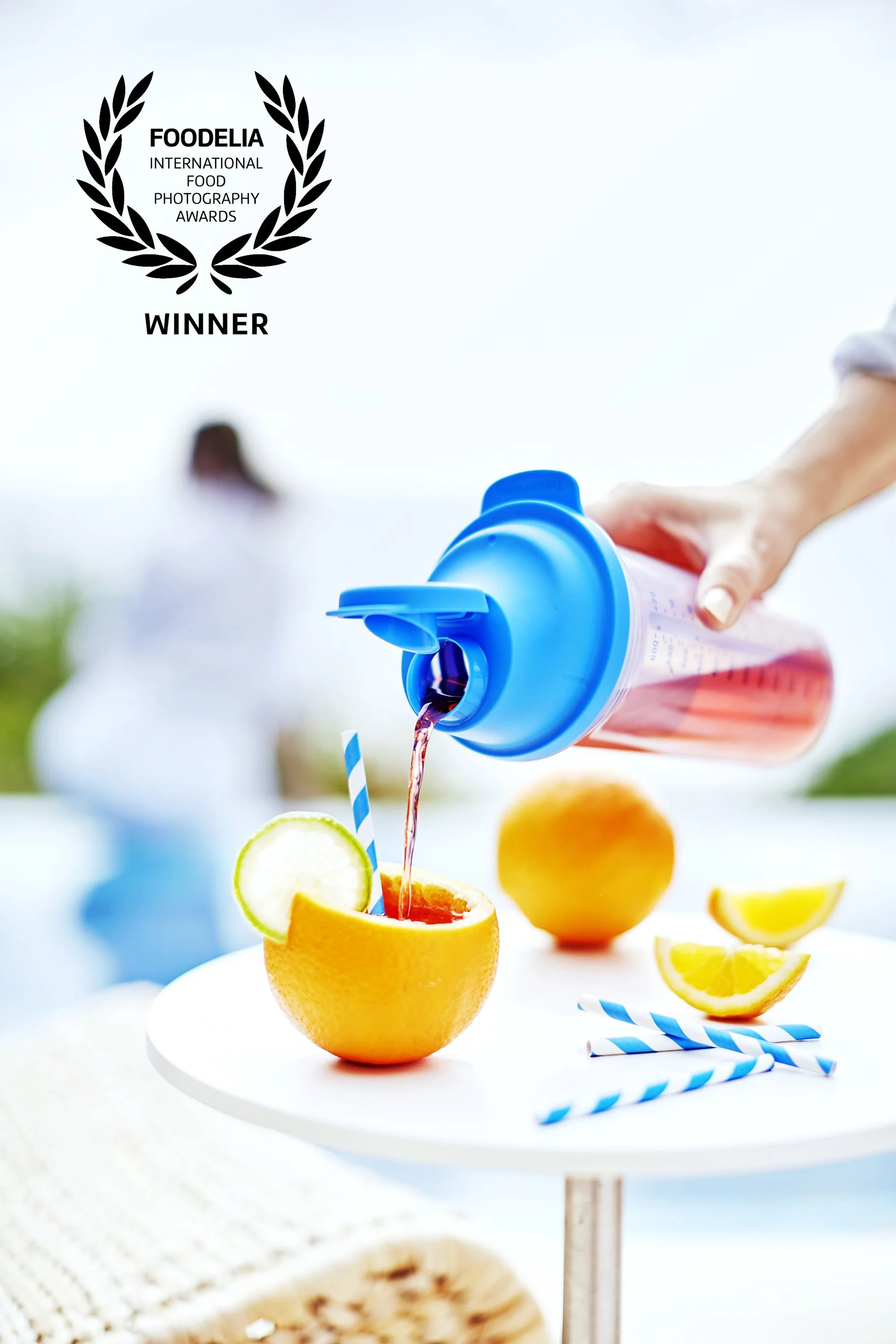 A person pouring pink liquid from a blue bottle into half an orange, which is used as a glass, on a white table. Lemon wedges and blue-striped straws are also on the table. A blurred person is in the background.