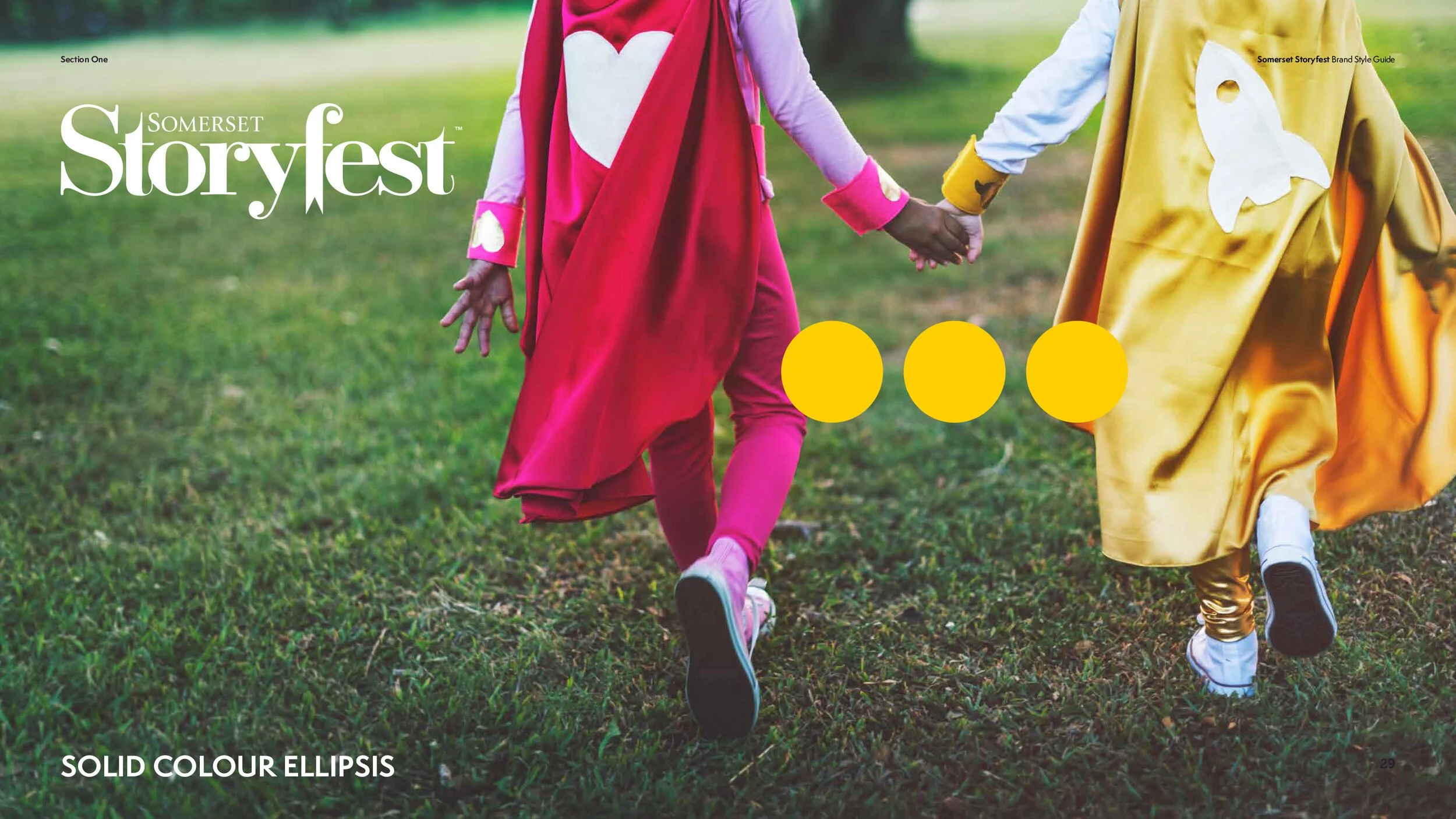 Two children wearing colorful superhero costumes holding hands and walking on a grassy field.