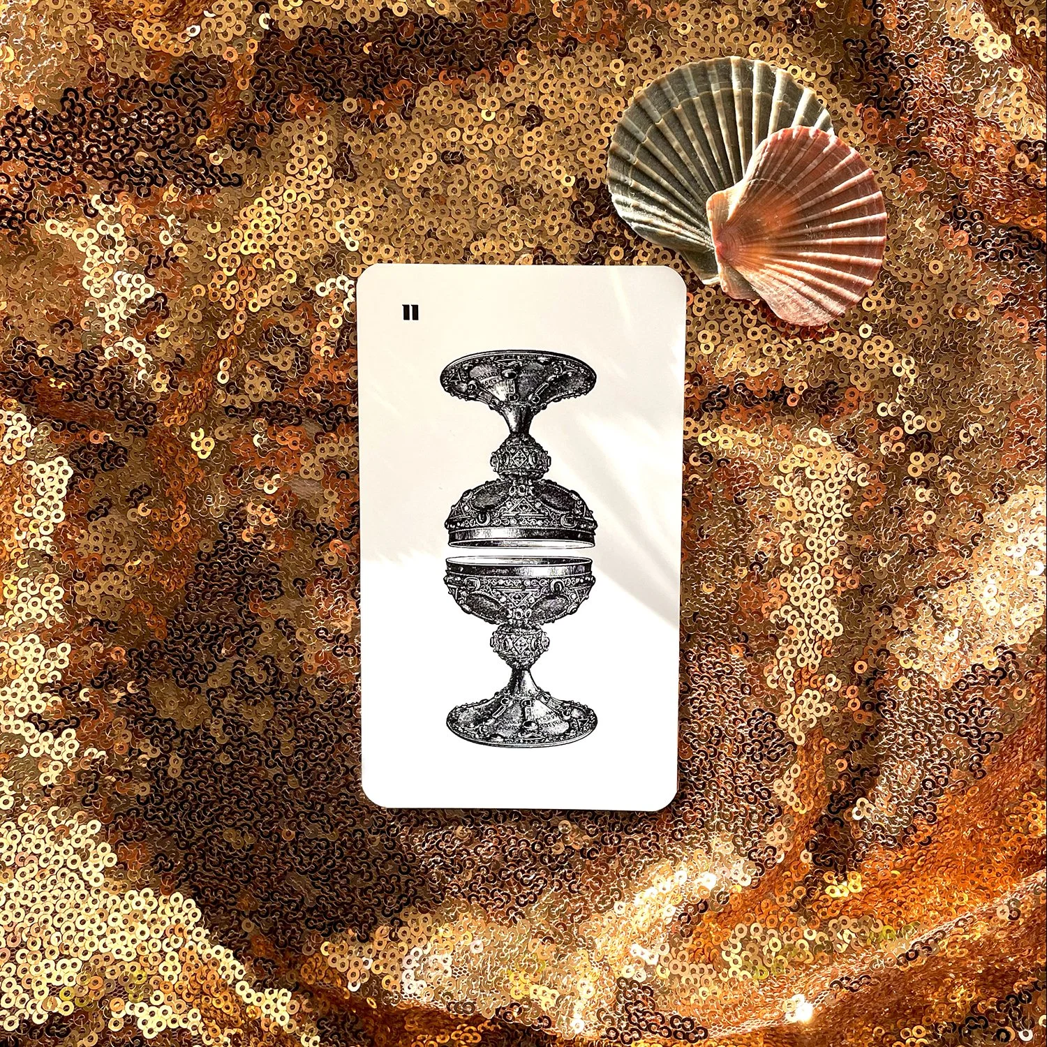 A tarot card with a black and white illustration of an ornate goblet or chalice, resting on a sequin-covered surface with two shells placed on the top right corner of the card.
