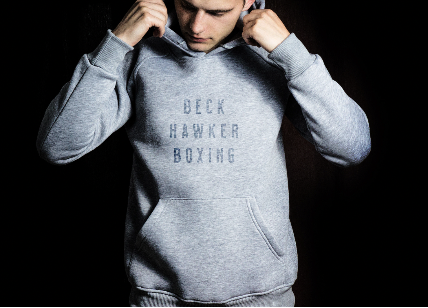 A young man puts on a gray hoodie with the words "BECK HAWKER BOXING" printed on the front, standing in front of a black background.