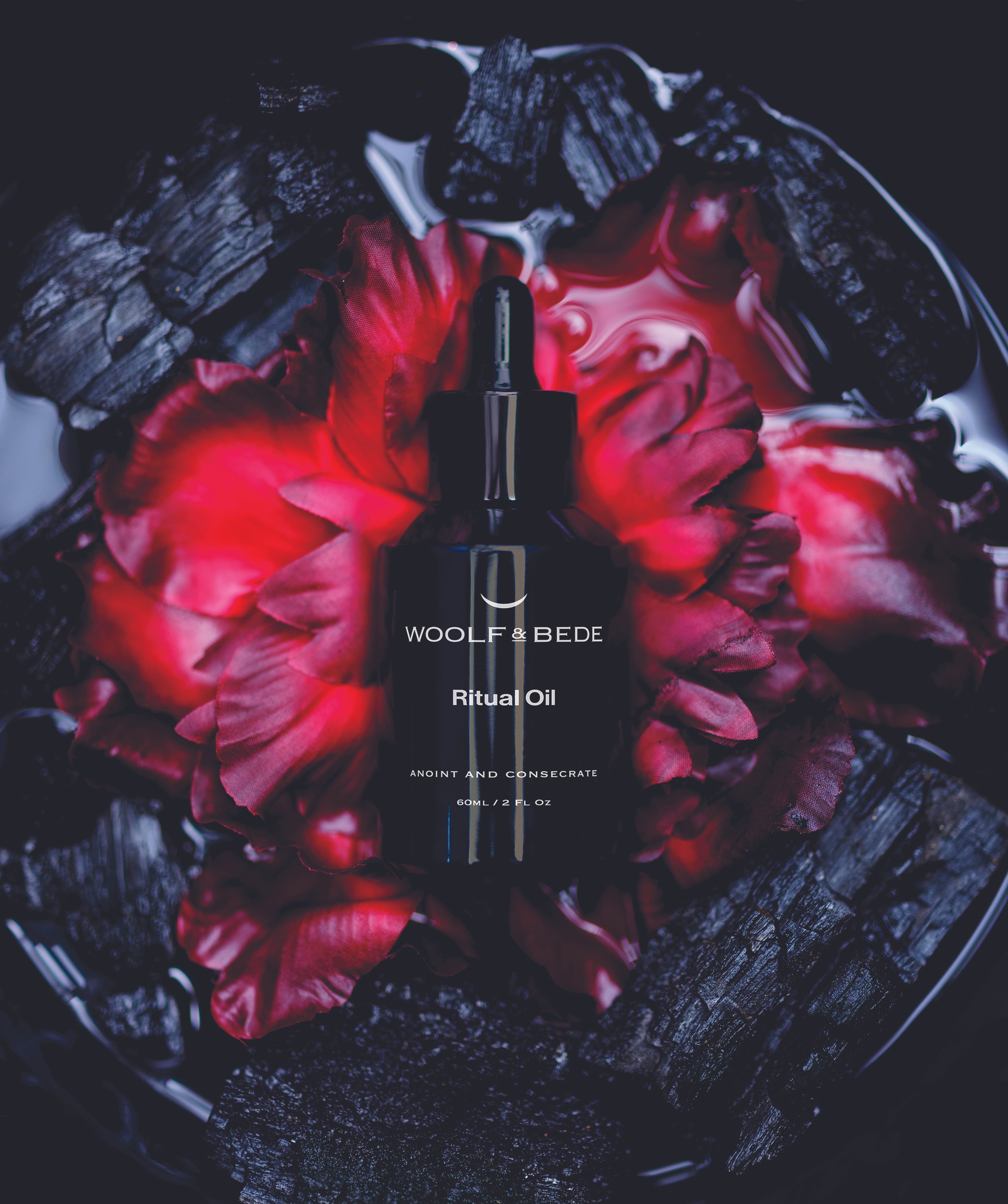A black bottle labeled 'Woolf & Bede Ritual Oil' is placed on red flower petals and surrounded by dark, charred wood pieces in a bowl.
