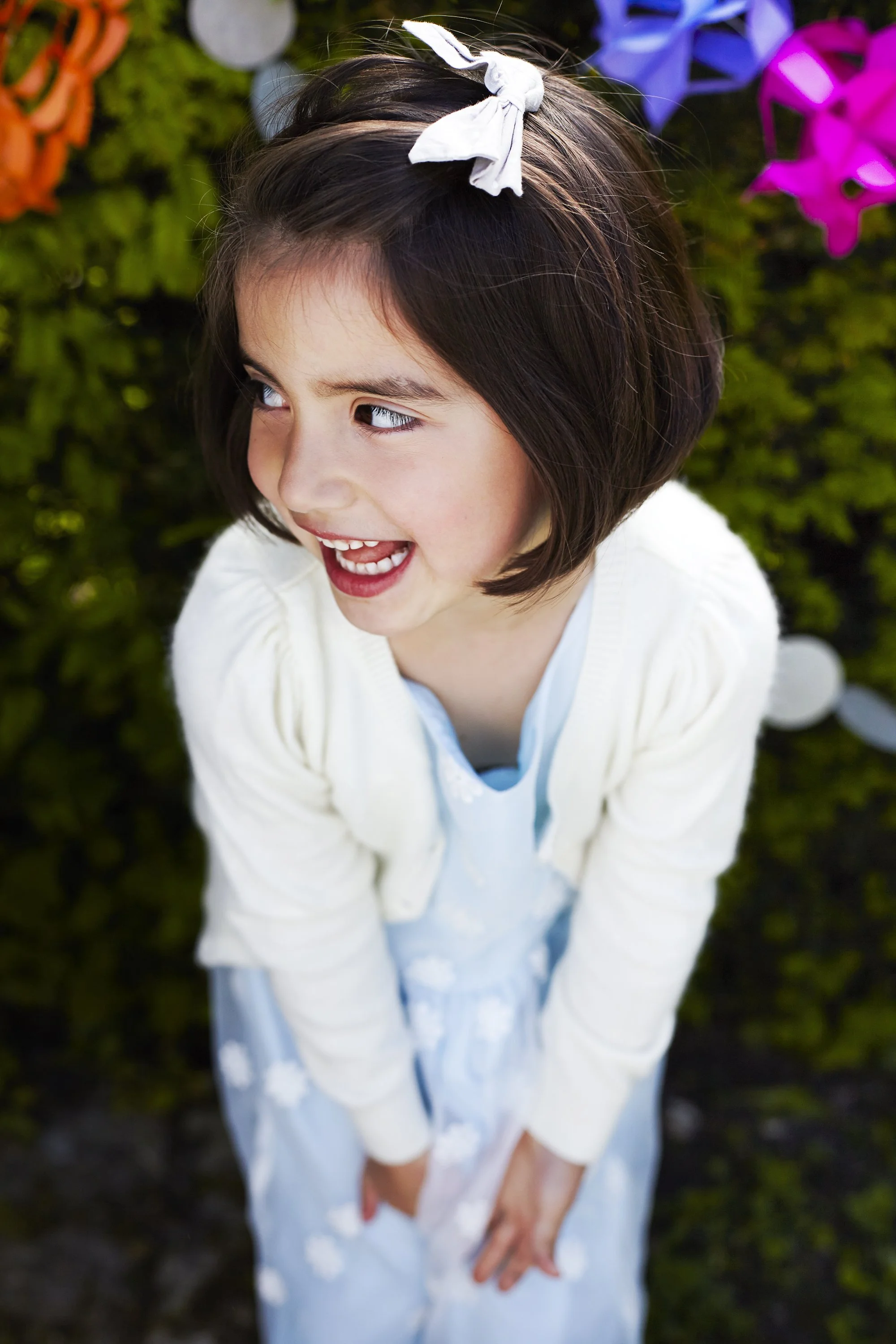 A young girl with short brown hair tied with a white bow, smiling and looking to her right, standing outdoors in front of colorful paper lanterns and greenery.