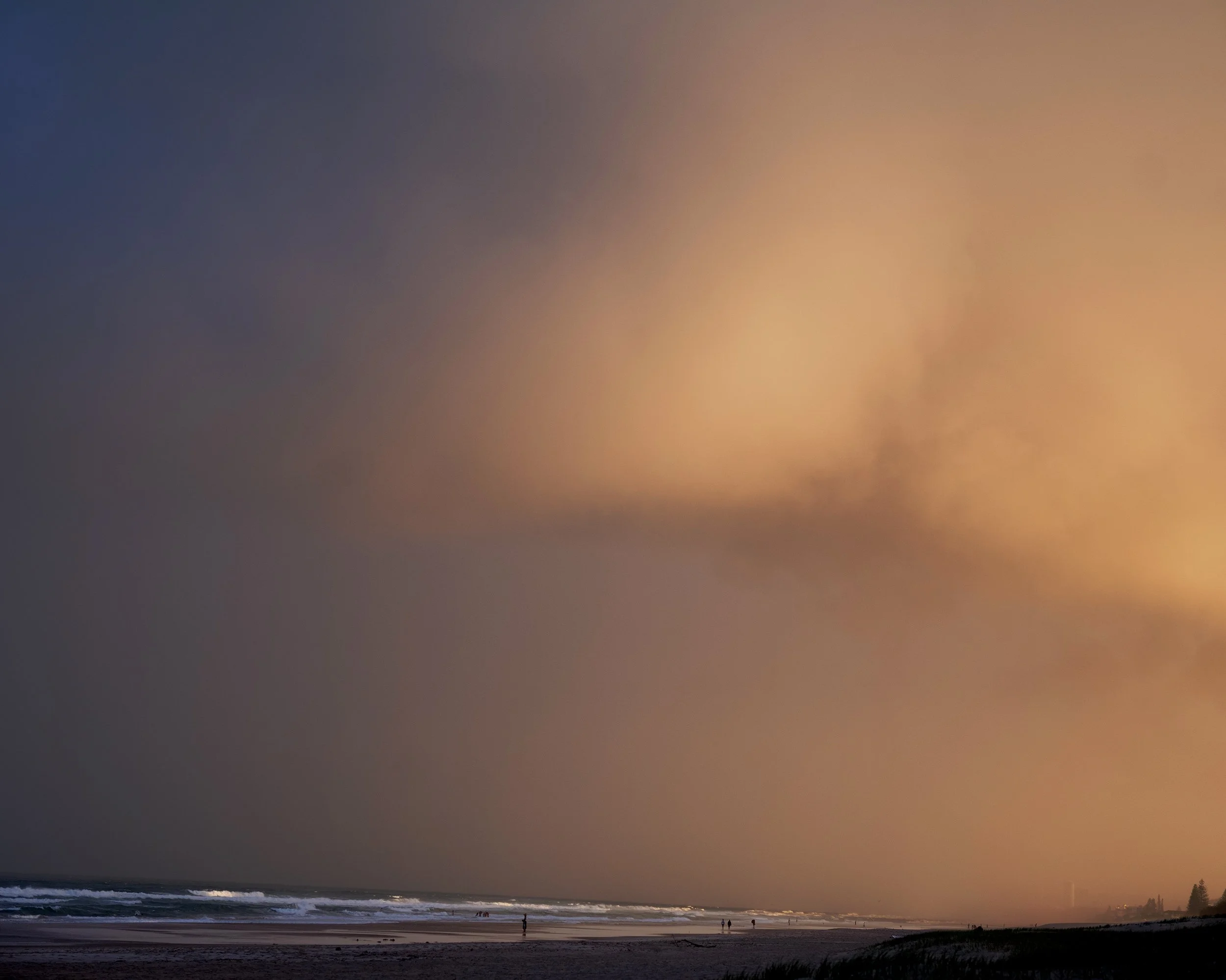 Beach at sunset with dark clouds and a rainbow forming in the sky. People are walking along the shoreline.