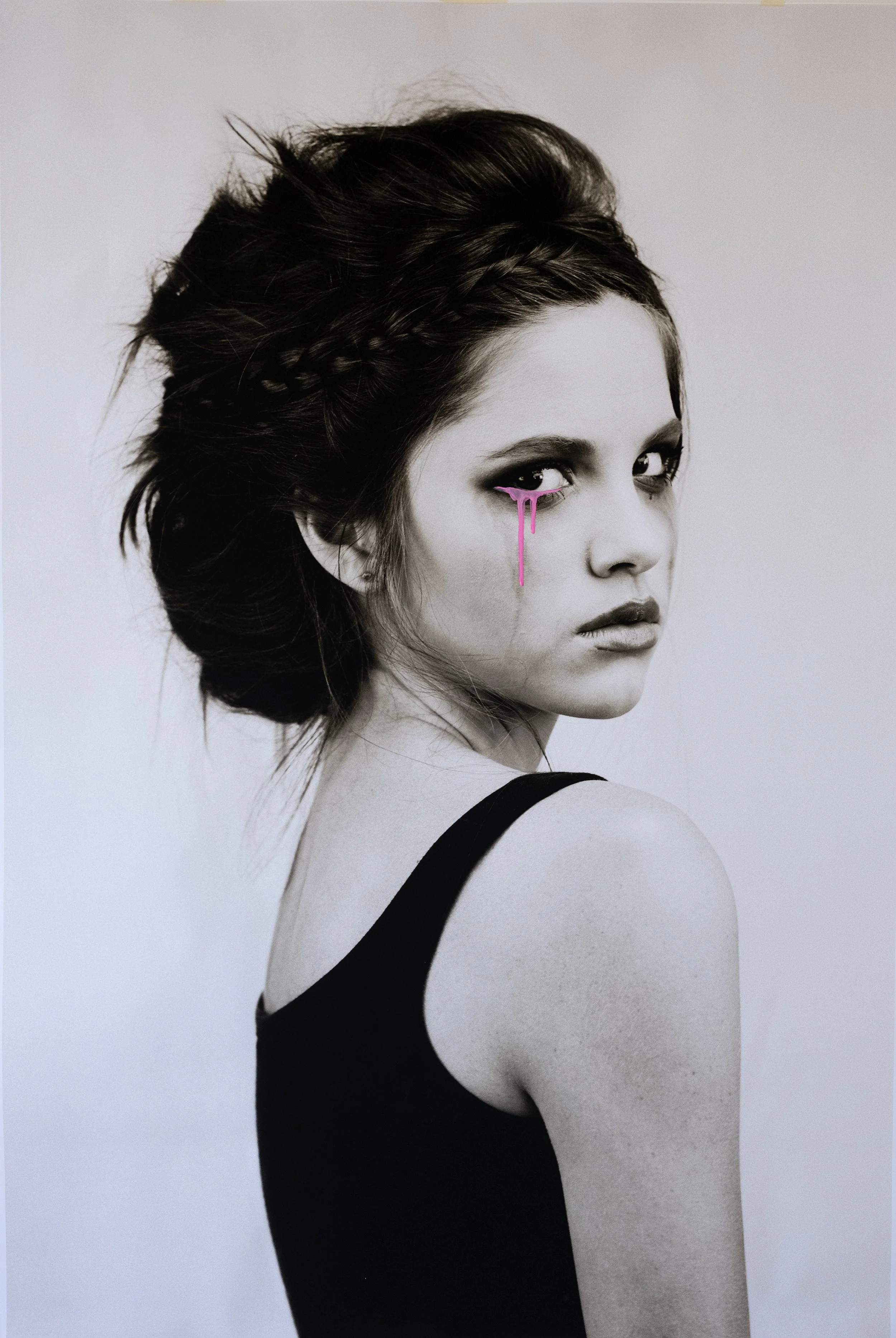 Black and white photograph of a woman with dark hair styled in a loose updo, wearing a black sleeveless top, with pink paint streaks running from her eyes.