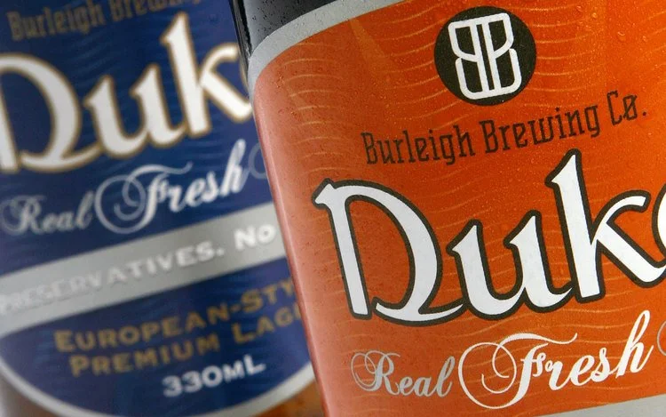 Close-up of two beer cans, one orange with 'Burleigh Brewing Co. Duke Real Fresh' and a black and white logo, and one blue label behind with similar branding.