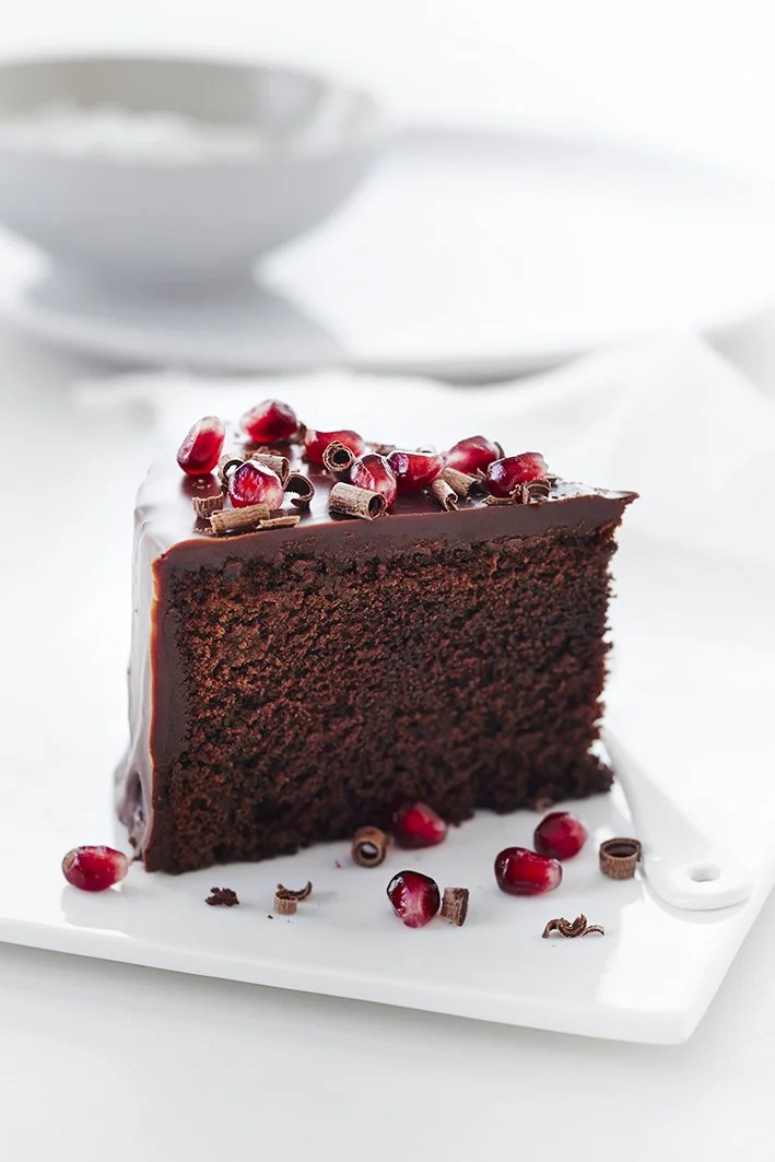 A slice of chocolate cake topped with pomegranate seeds and chocolate curls on a white cutting board.