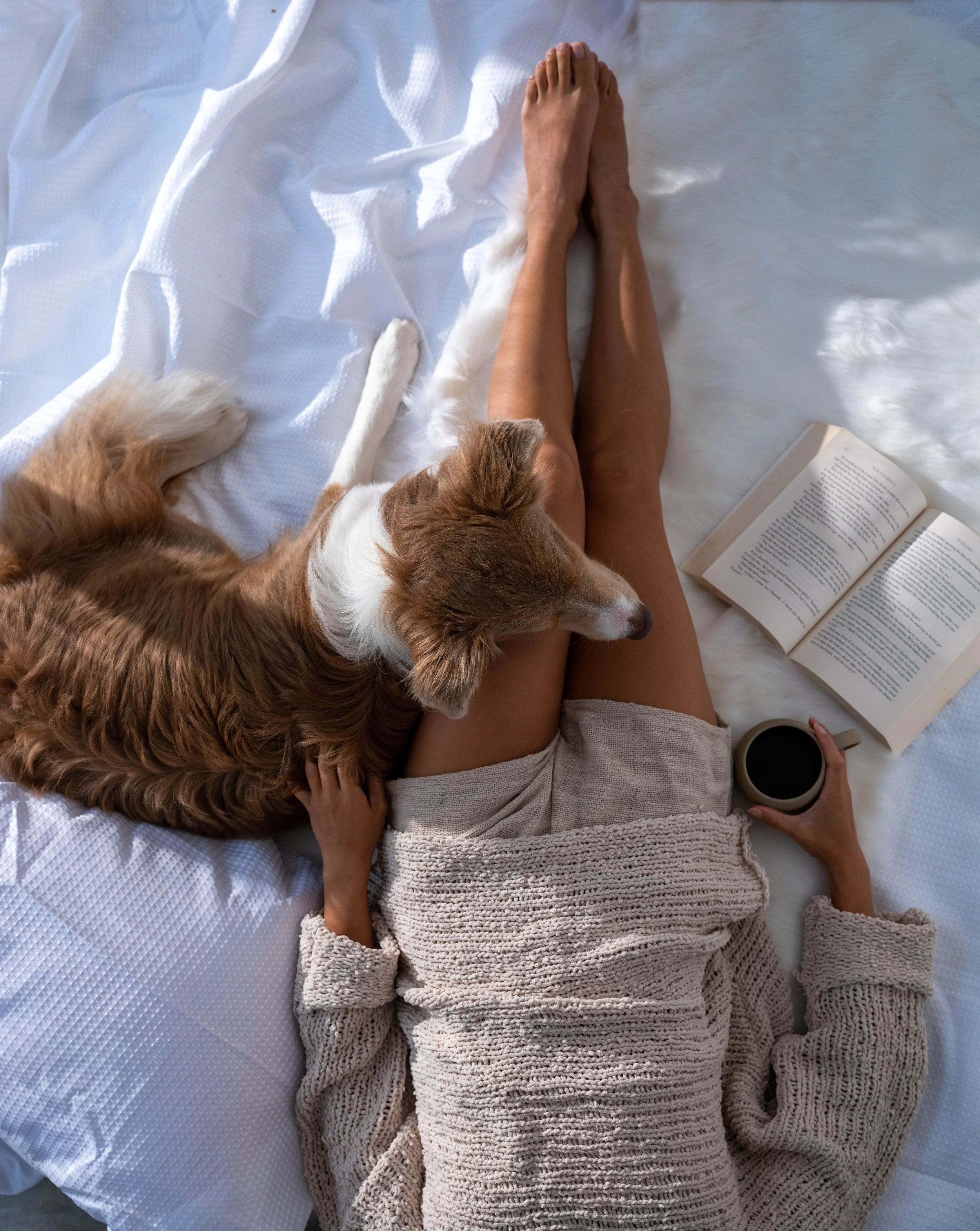 A person lying on a bed with a dog laying on their side, a book open nearby, and a mug of coffee or tea in their hand.