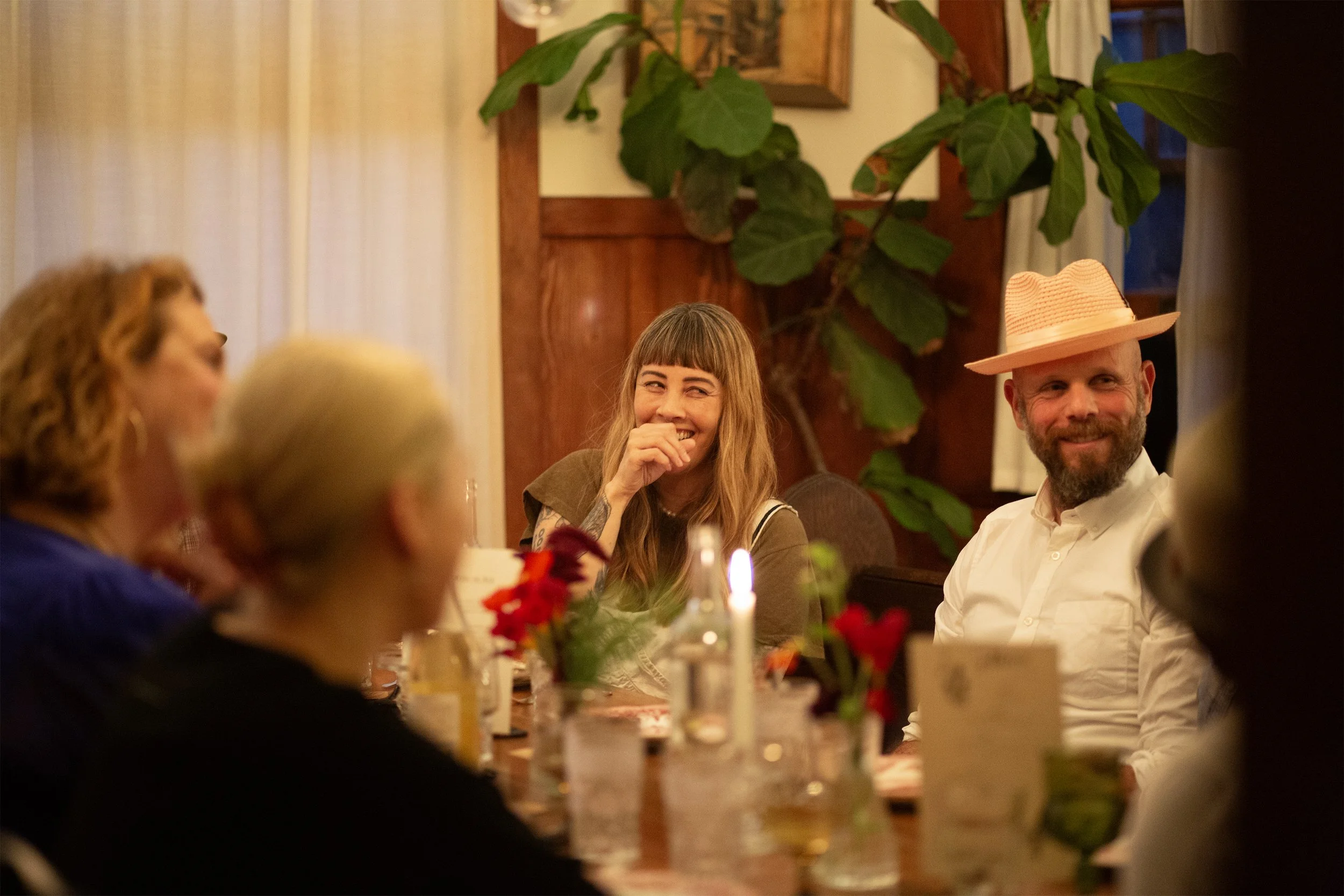 A group of people sitting around a table at a dinner gathering, smiling and laughing, with a woman in the center wearing a brown top and a man on the right wearing a white shirt and a straw hat.