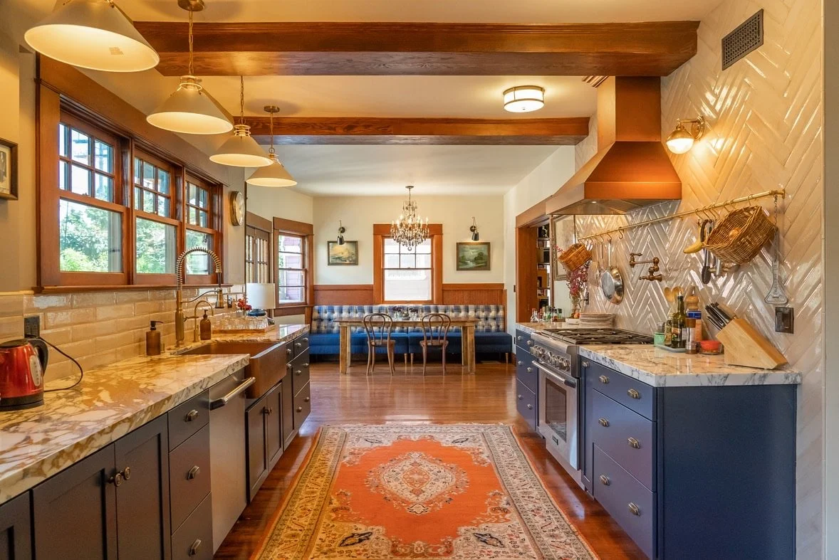Kitchen with dark blue lower cabinets, marble countertops, a row of windows to the left, and a dining area with a built-in blue booth and wooden chairs at the back. The kitchen has wooden beams, a chandelier over the dining table, and a rug with oran