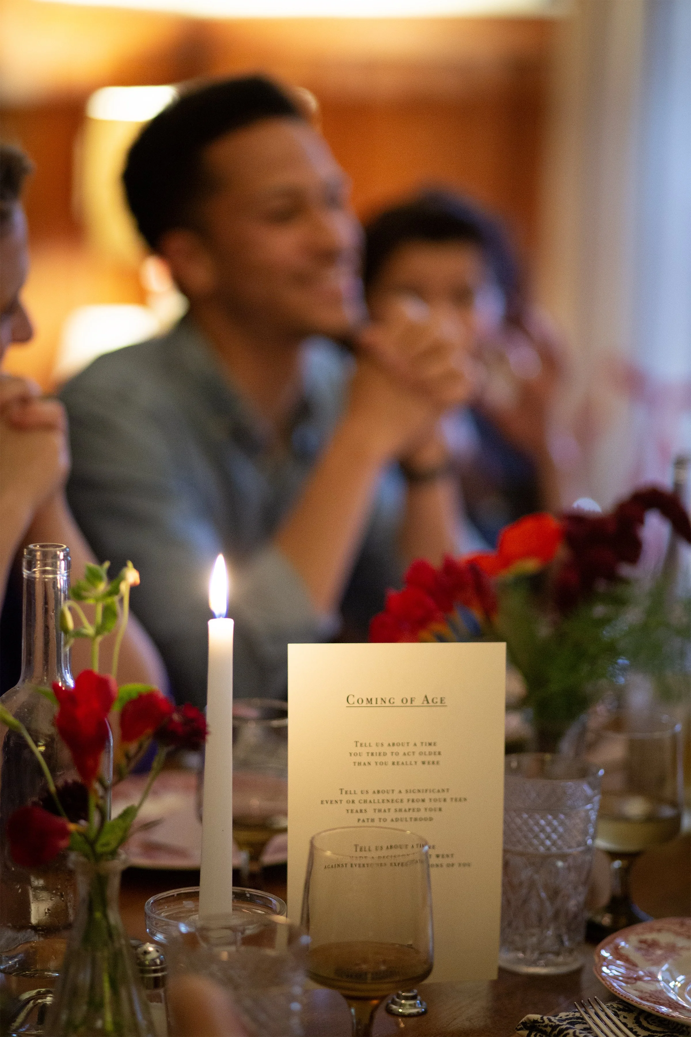 A dining table with flowers, candles, drinks, and a menu, with people smiling and talking in the background at a gathering.