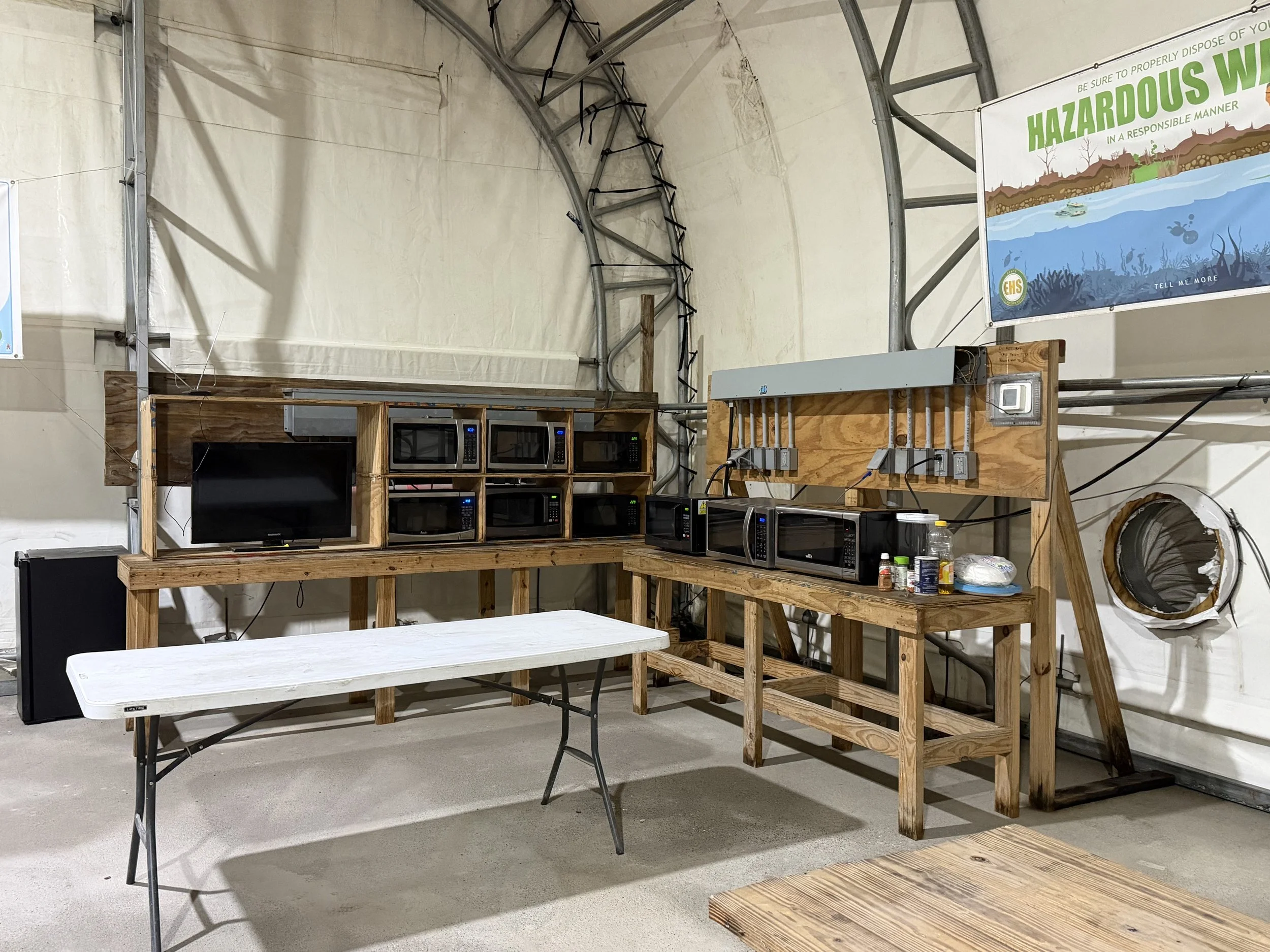 Interior of a greenhouse or workshop with wooden shelving holding multiple microwaves and a TV, a white folding table, and electric outlets on a wood panel, with posters about hazardous waste on the walls.Janitorial Services