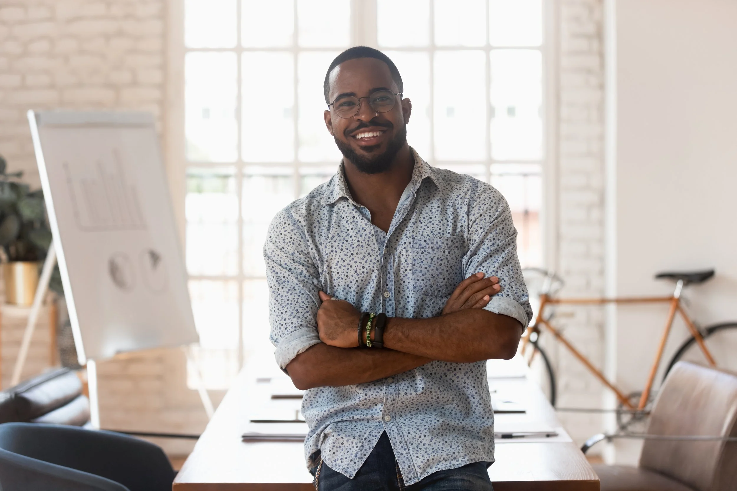 A smiling man with glasses and a short beard stands with arms crossed in a bright, modern office space with large windows, a whiteboard, books, a bicycle, and a wooden table.