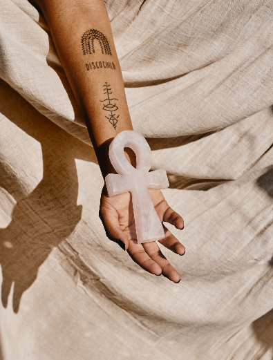 Person holding a white anchor-shaped object, dressed in beige fabric, with tattoos on their arm.
