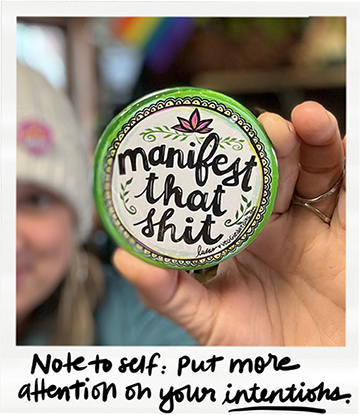A person holding a round container with a label that reads "manifest that shit." The photo includes a handwritten note saying, "Note to self: Put more attention on your intentions."