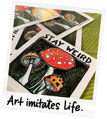 A stack of illustrated cards with mushroom designs, one of which has a message that says "STAY WEIRD." The bottom card has a quote that says "Art imitates life." The cards are placed on a green surface.