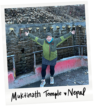 Person smiling and raising arms in front of a wall with hanging bells at Muktinath Temple in Nepal.