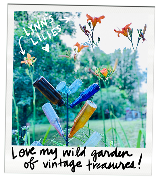 Colorful glass bottles arranged on sticks in a vibrant garden with blooming lilies labeled "Lynn's lilies", and text saying "Love my wild garden of vintage treasures!"