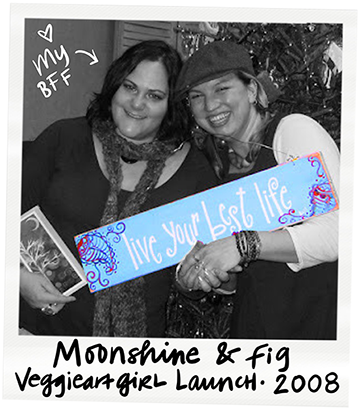 Two women smiling and holding a blue sign that says 'Live your best life' at a 2008 veggie girl launch event themed 'Moonshine & Fig,' standing in front of a decorated background.