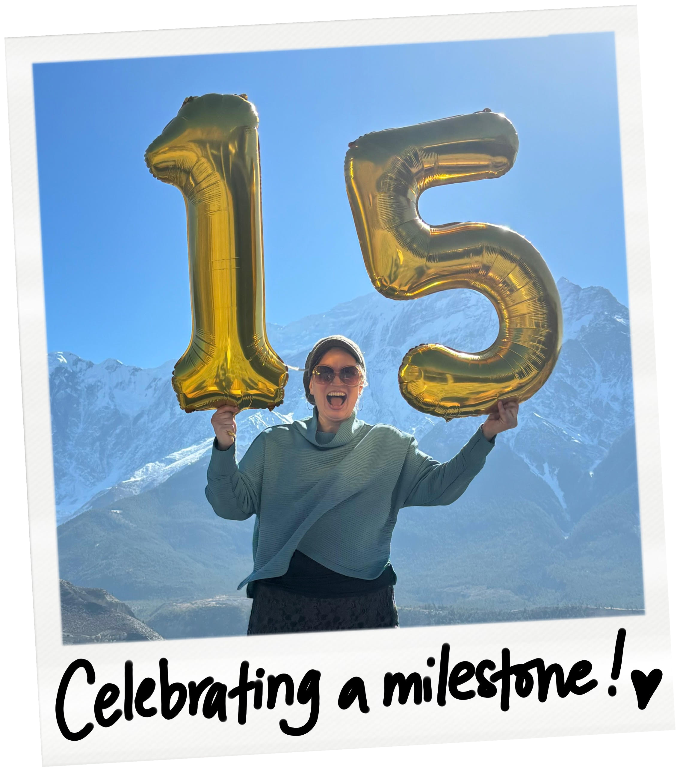 A woman holding large gold balloons shaped as the numbers 1 and 5, celebrating a milestone with snow-capped mountains in the background and a handwritten message that says "Celebrating a milestone!"