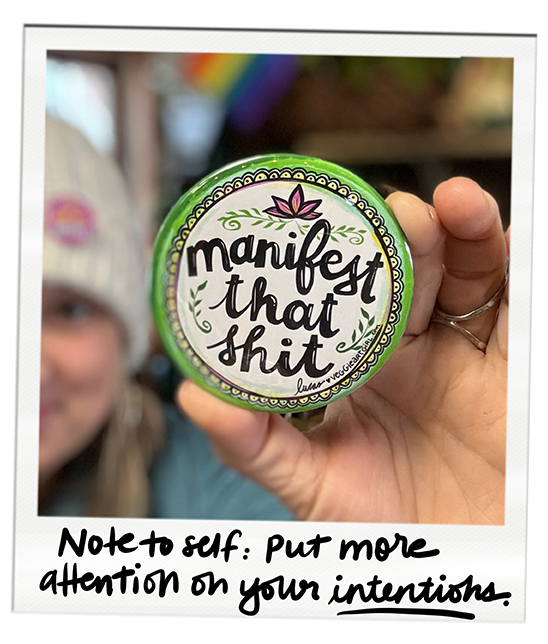 Person holding a round green container with a label that reads "manifest that shit" in black handwritten font, featuring a pink lotus flower and decorative green designs. The background is blurry with a hint of rainbow colors.