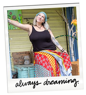 Woman with blonde hair, tattoos, wearing a black tank top, blue headscarf, and colorful skirt, smiling with arms outstretched outdoors on a porch.