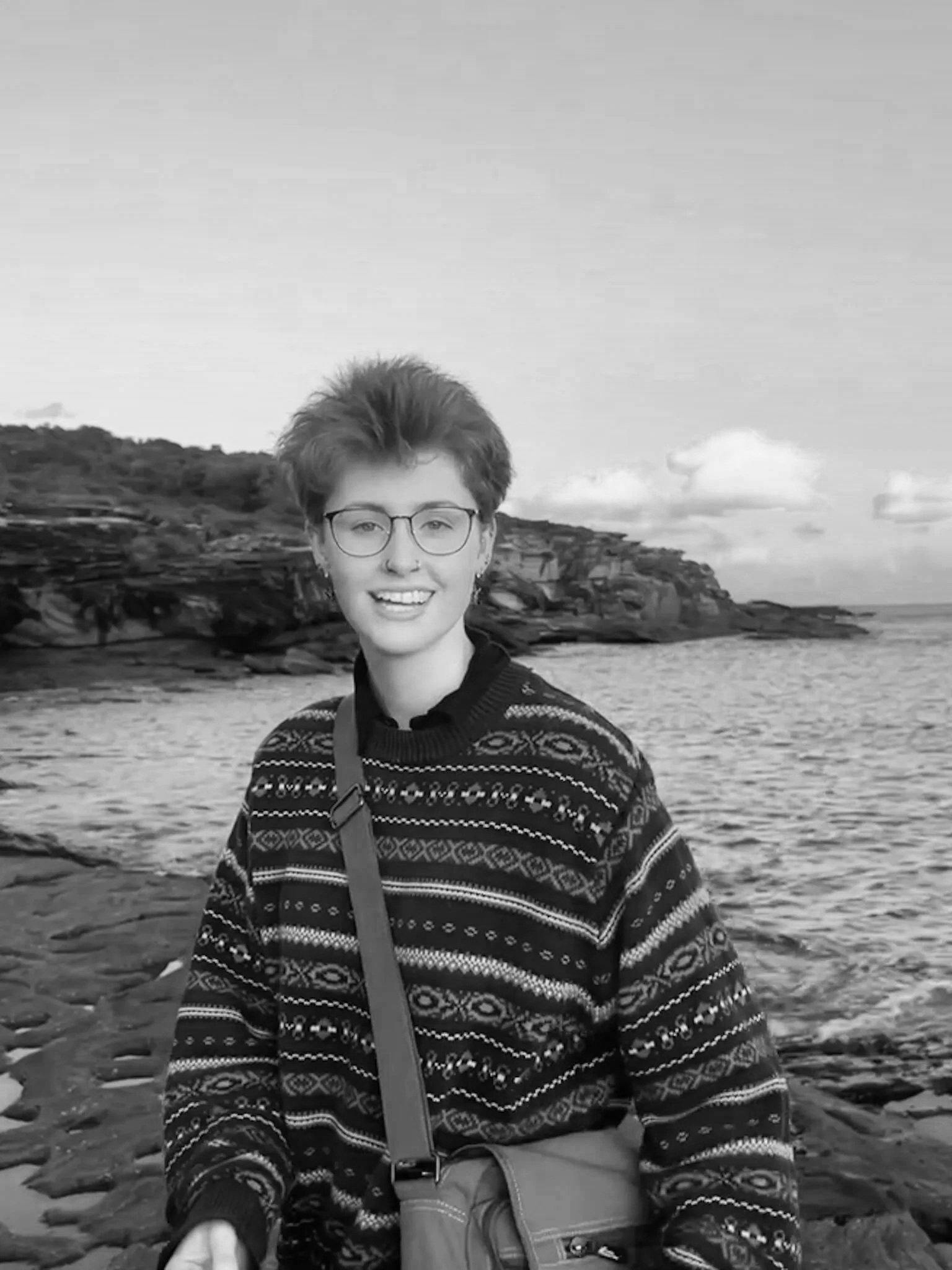 A smiling young person with glasses and short hair, wearing a patterned sweater, standing on a rocky shoreline with water and cliffs in the background.