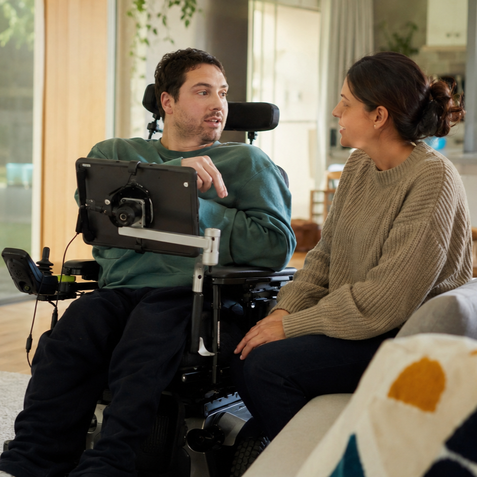 A man in a wheelchair with a tablet talking to a woman sitting on a couch, both smiling and engaged in a conversation in a cozy, well-lit living room.