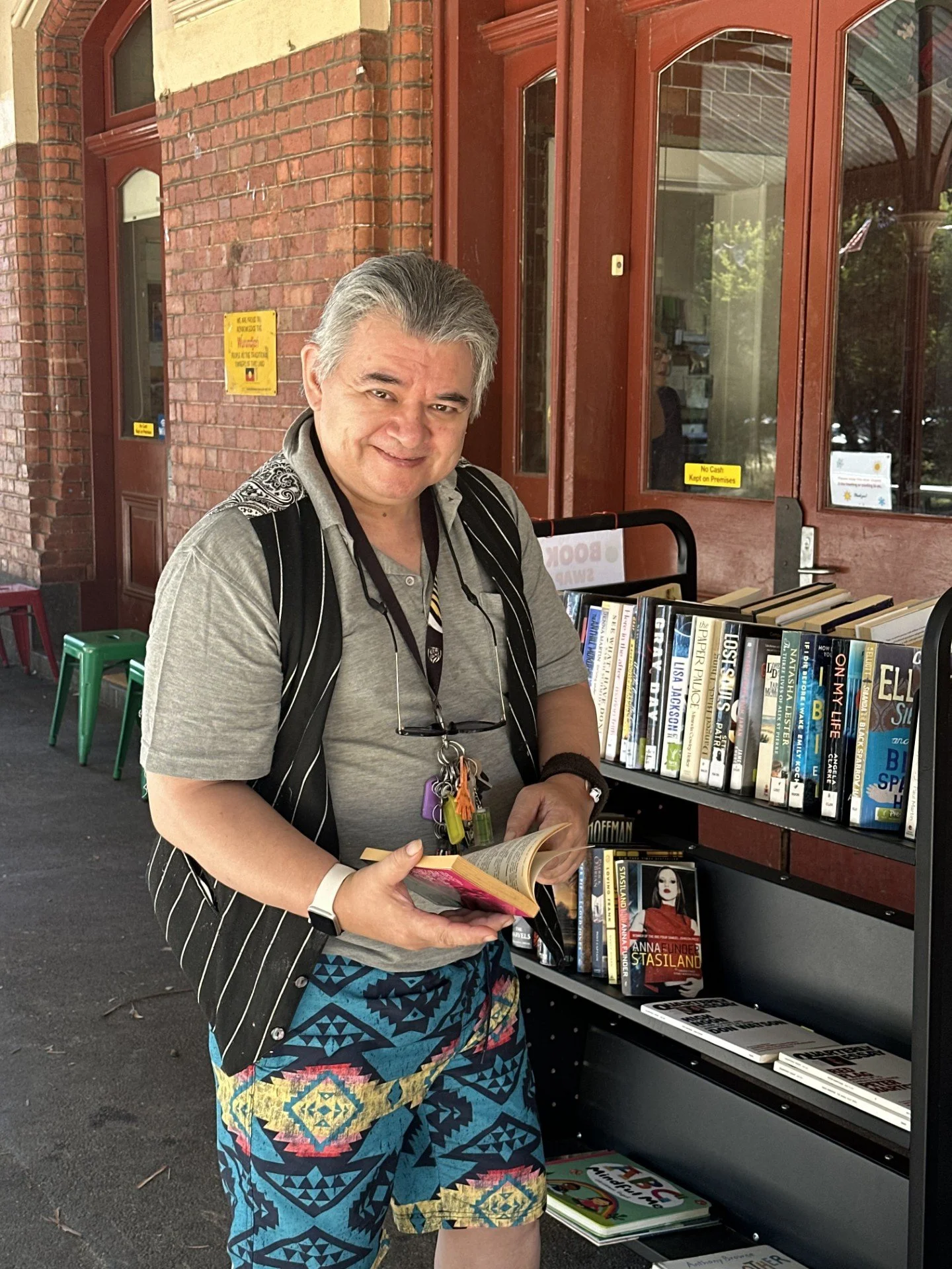 A man with gray hair wearing a striped vest, a gray shirt, and colorful shorts stands outside a bookstore, looking at a book in his hands. Behind him are books on a shelf and a brick building with large windows. He has a lanyard around his neck and keys hanging from it.
