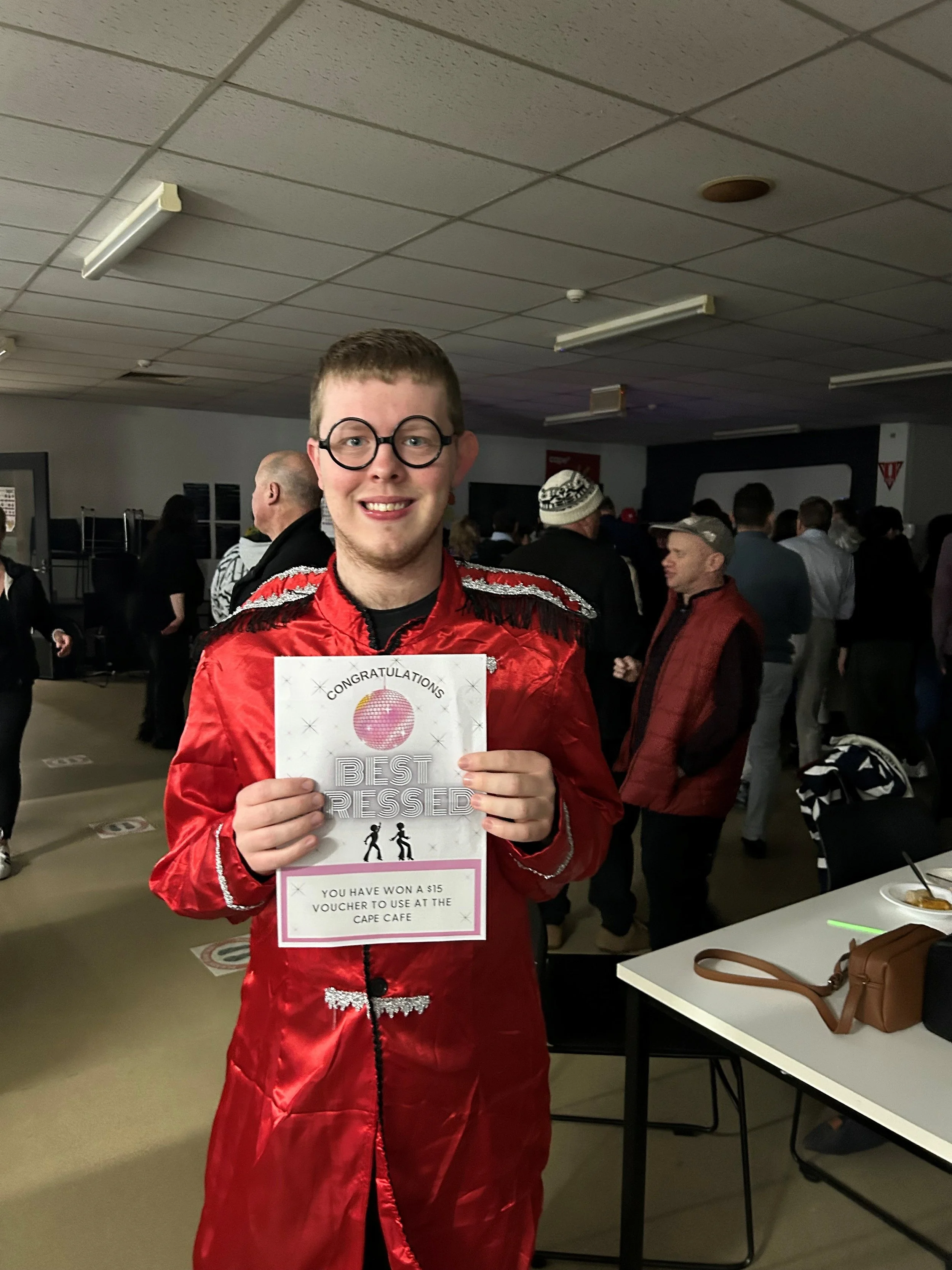 A young man in a red satin jacket and glasses holding a certificate that says 'Congratulations Best Dressed' at an indoor event with other people in the background.