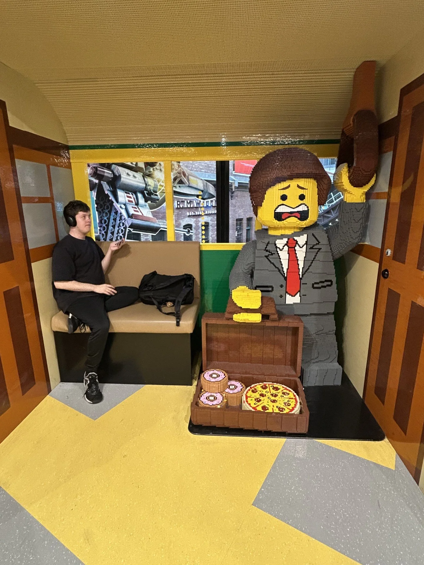 A person sitting on a beige bench wearing headphones and holding an object, next to a LEGO sculpture of a man in a suit holding a phone, with LEGO pizza and donuts on a tray in front.