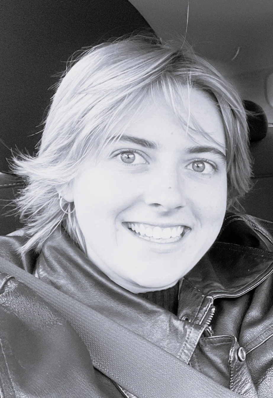 Alex, Support Worker. A black and white close-up photo of a smiling woman with short, layered hair wearing a leather jacket and a seatbelt.