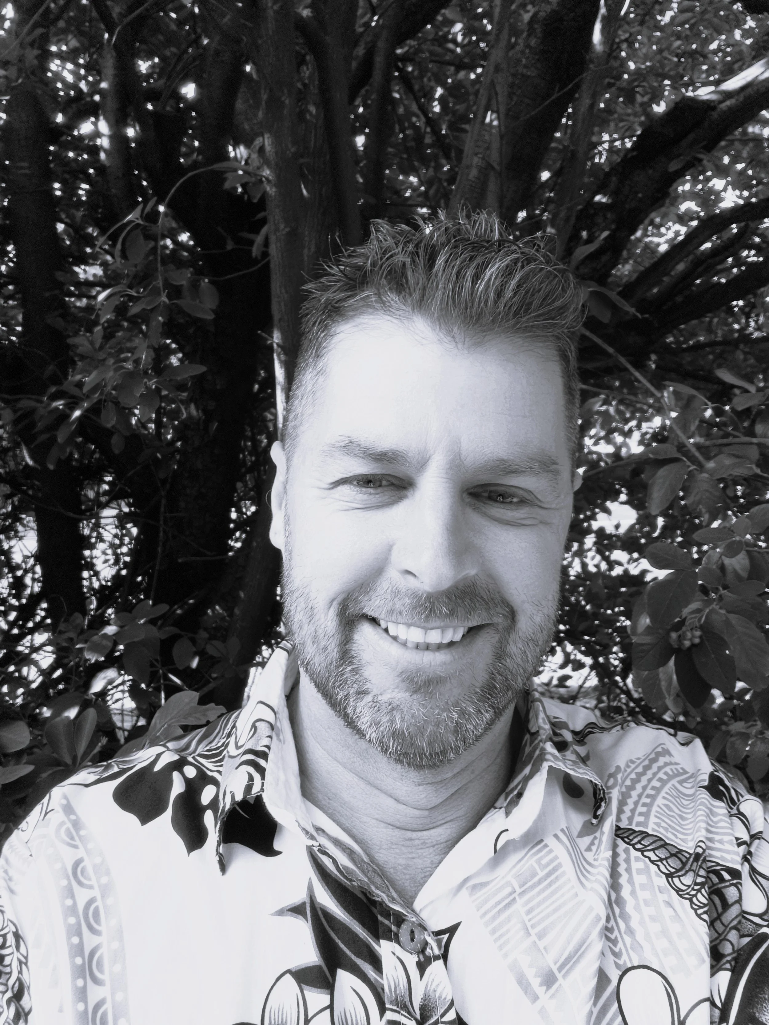 Ben, Support Worker. Black and white photo of a smiling man with short, spiked hair and a beard, wearing a patterned Hawaiian shirt, standing in front of a dense tree with leaves and branches.