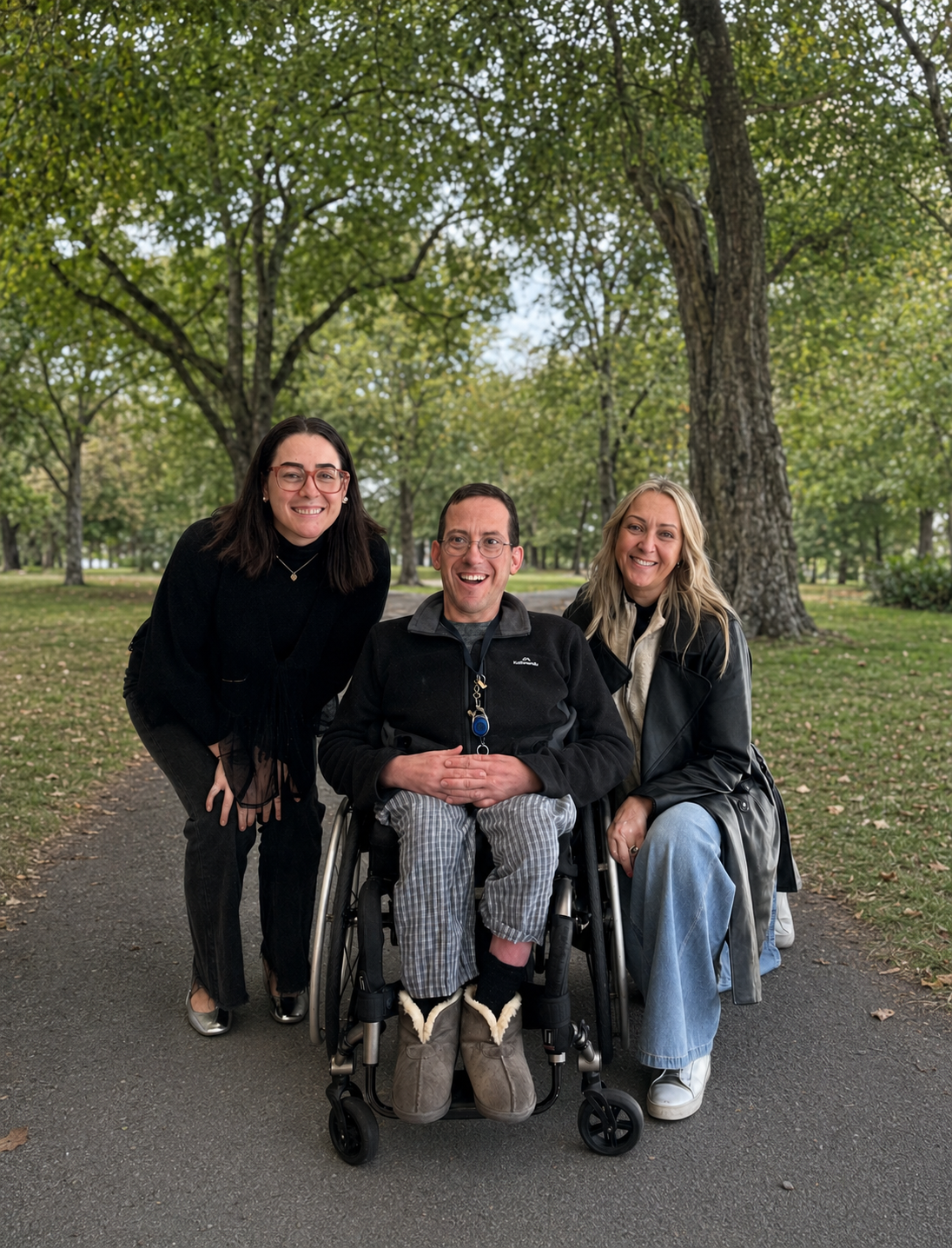 Three smiling adults pose together on a paved path in a leafy park. A man using a wheelchair sits between two women, surrounded by mature trees and grass.