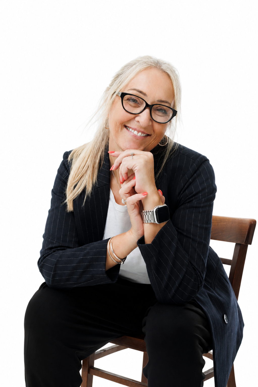 A smiling middle-aged woman with blonde hair and glasses, wearing a black pinstripe blazer, white top, and black pants, sitting on a wooden chair against a white background, with her chin resting on her clasped hands.