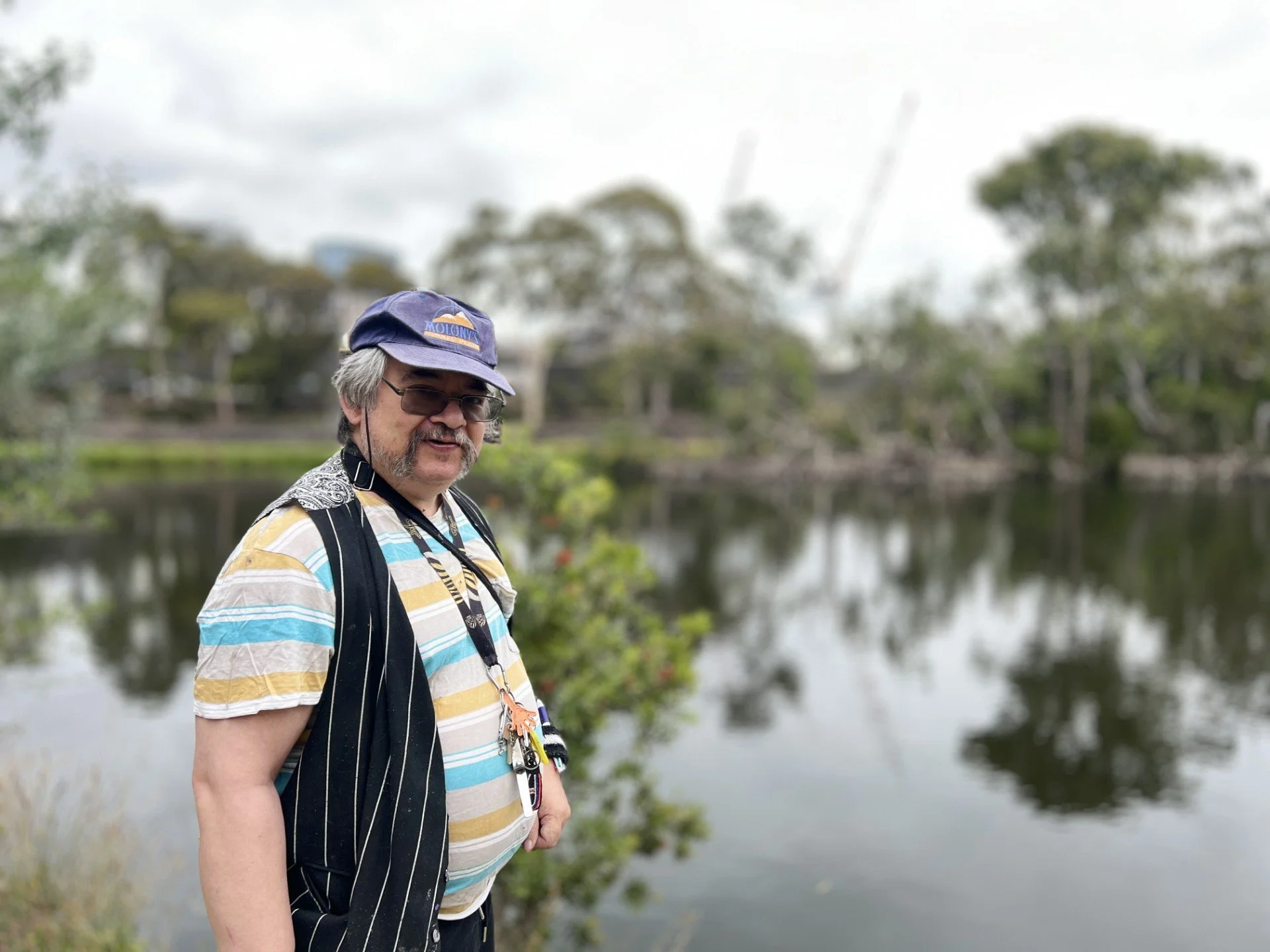 A man with gray hair and a mustache wearing sunglasses, a blue cap, a colorful striped shirt, and a camera strap around his neck stands outdoors by a body of water with trees in the background on an overcast day.