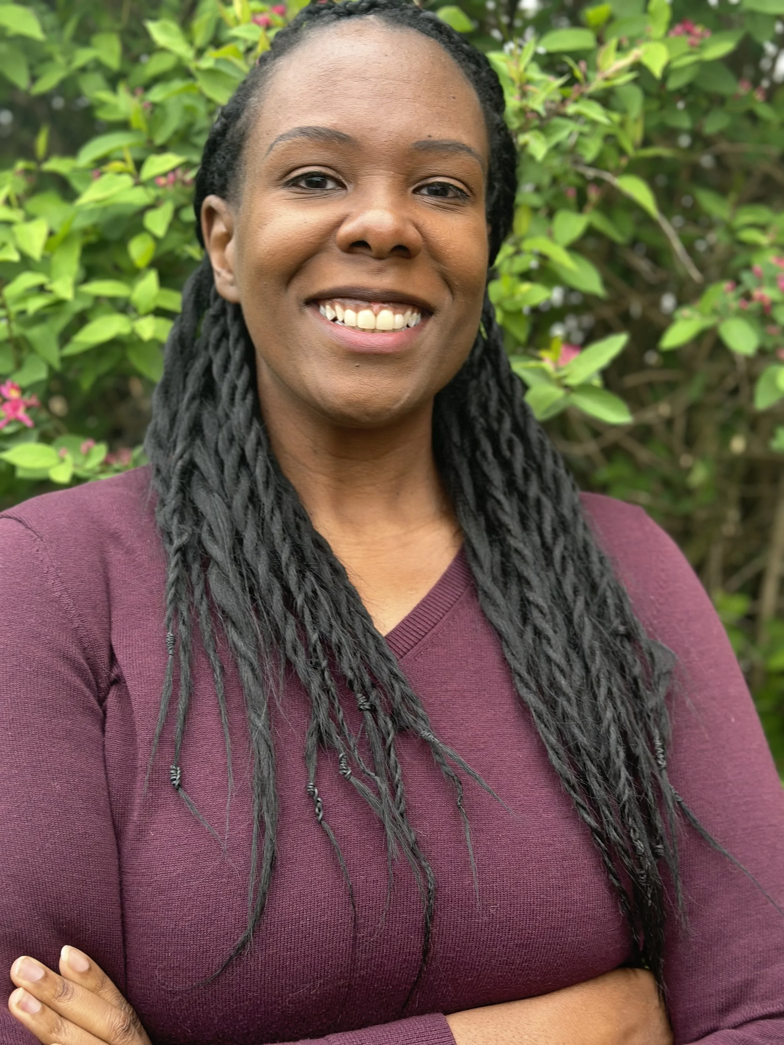 Portrait of a smiling woman with long twisted hair, wearing a maroon long-sleeve top, standing outdoors in front of green foliage and pink flowers.