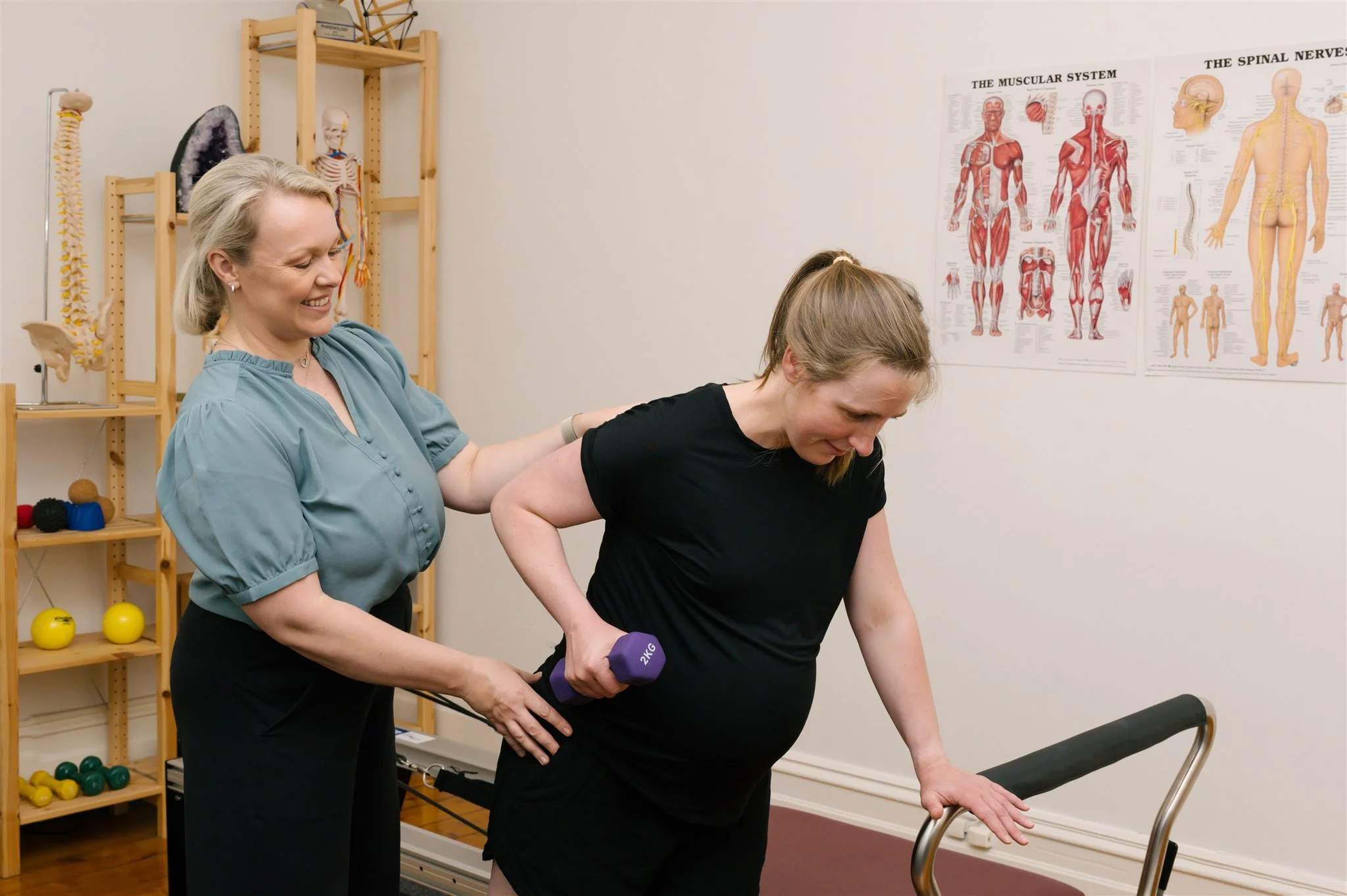 A physical therapist helps a woman with a black shirt and black shorts perform a back exercise with a purple dumbbell in a rehab room. The room has anatomical posters on the wall and shelves with colorful therapy tools.