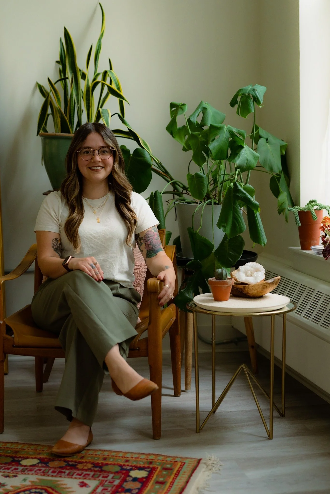 A woman with glasses, long wavy hair, and tattoos on her arm, sitting on a wooden chair in a room with houseplants, a colorful rug, and a small table with decorative items and plants.