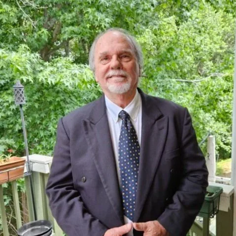 Close-up of an elderly man with grey hair and freckles, wearing a checkered shirt and a blue T-shirt underneath, standing outdoors with a blurred green leafy background.