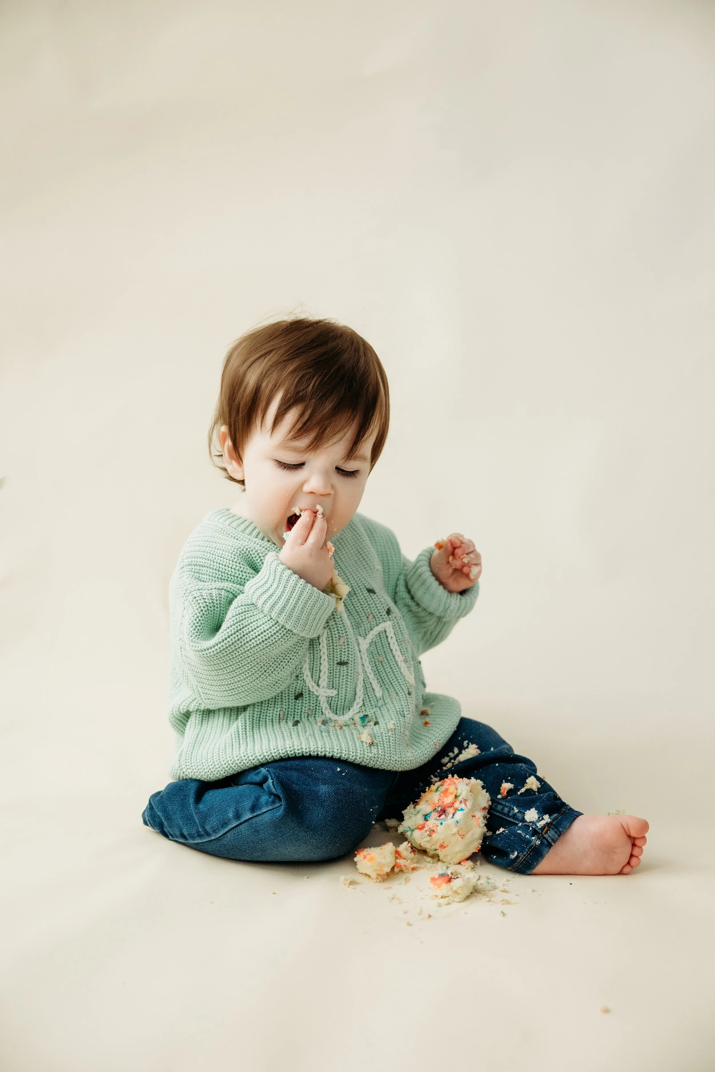 One-year-old toddler enjoying a colorful cake smash during a first birthday photography session with a neutral studio backdrop in Fort Myers, Florida.