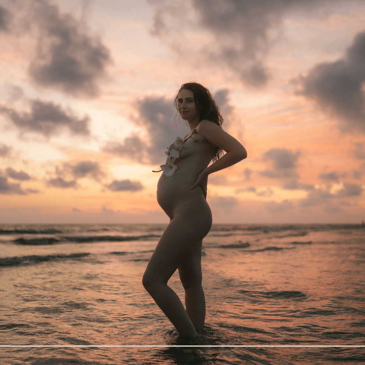 pregnancy mom is standing nude in the water during golden hour with an orchid covering her breasts during a maternity photography session on fort myers beach florida