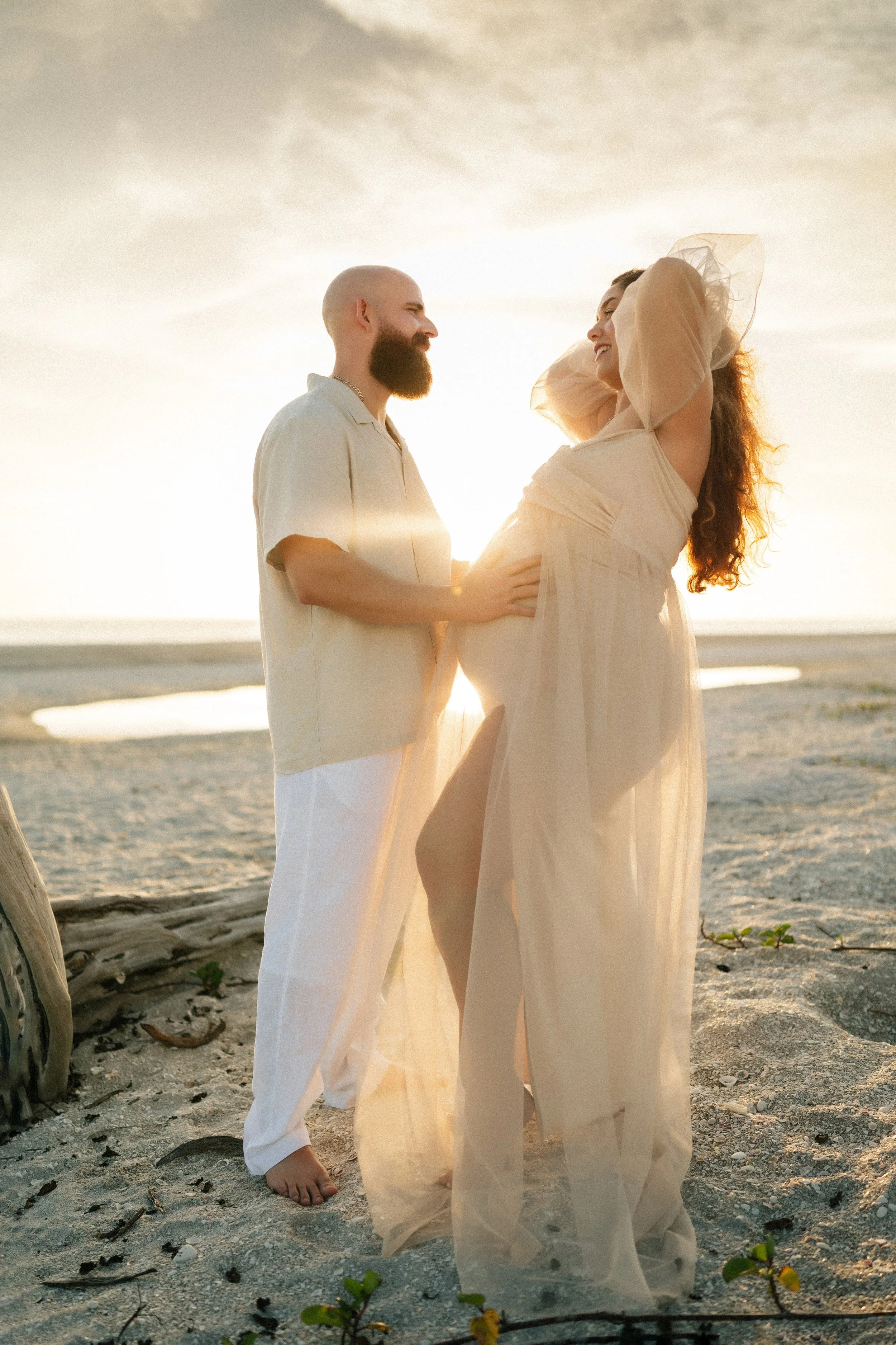 Expecting parents embracing on the beach during a golden hour maternity photography session with flowing dress and sunset light on Sanibel Island, Florida.
