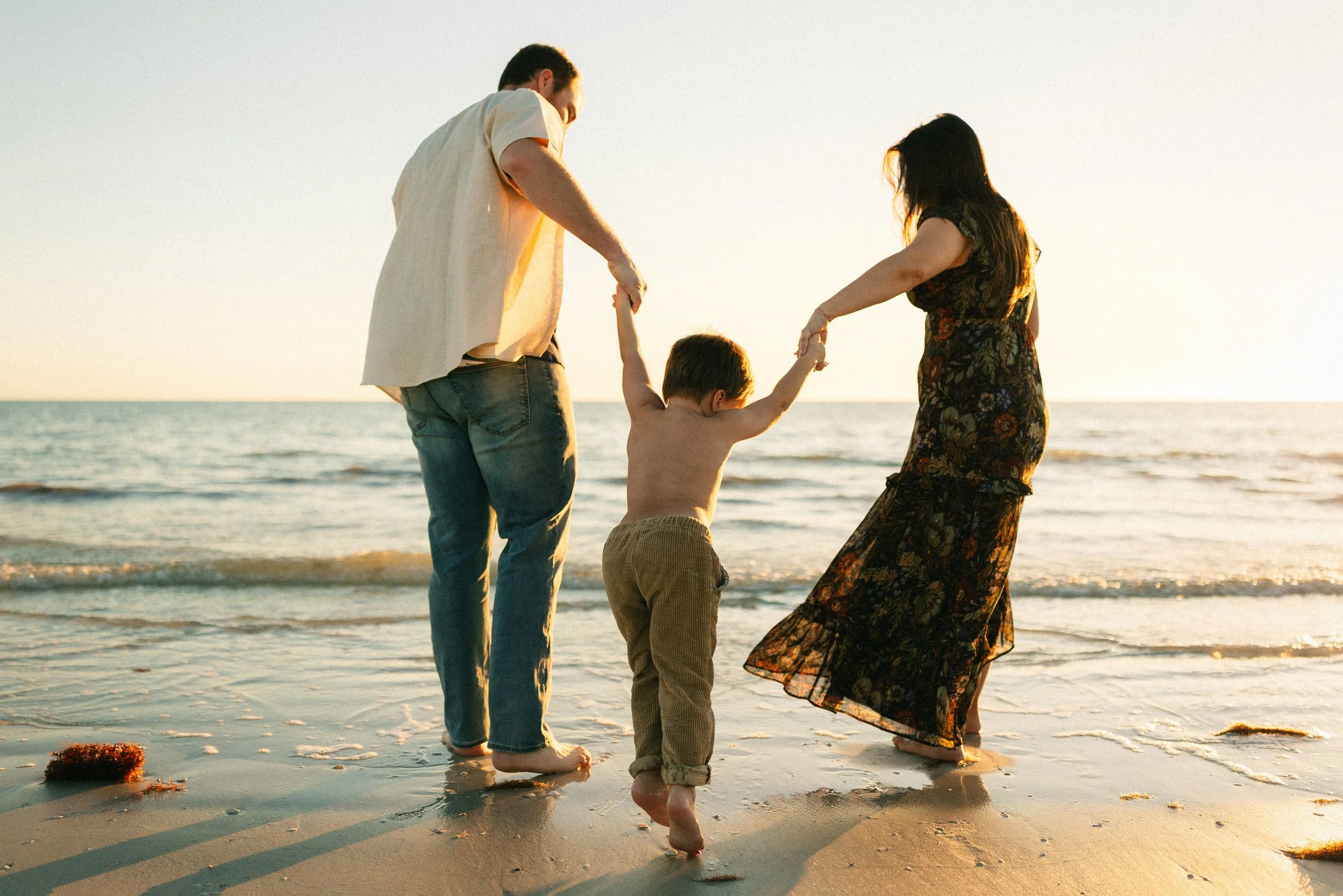 Parents holding their young child's hands while walking along the shoreline during a sunset family photography session on a Southwest Florida beach.
