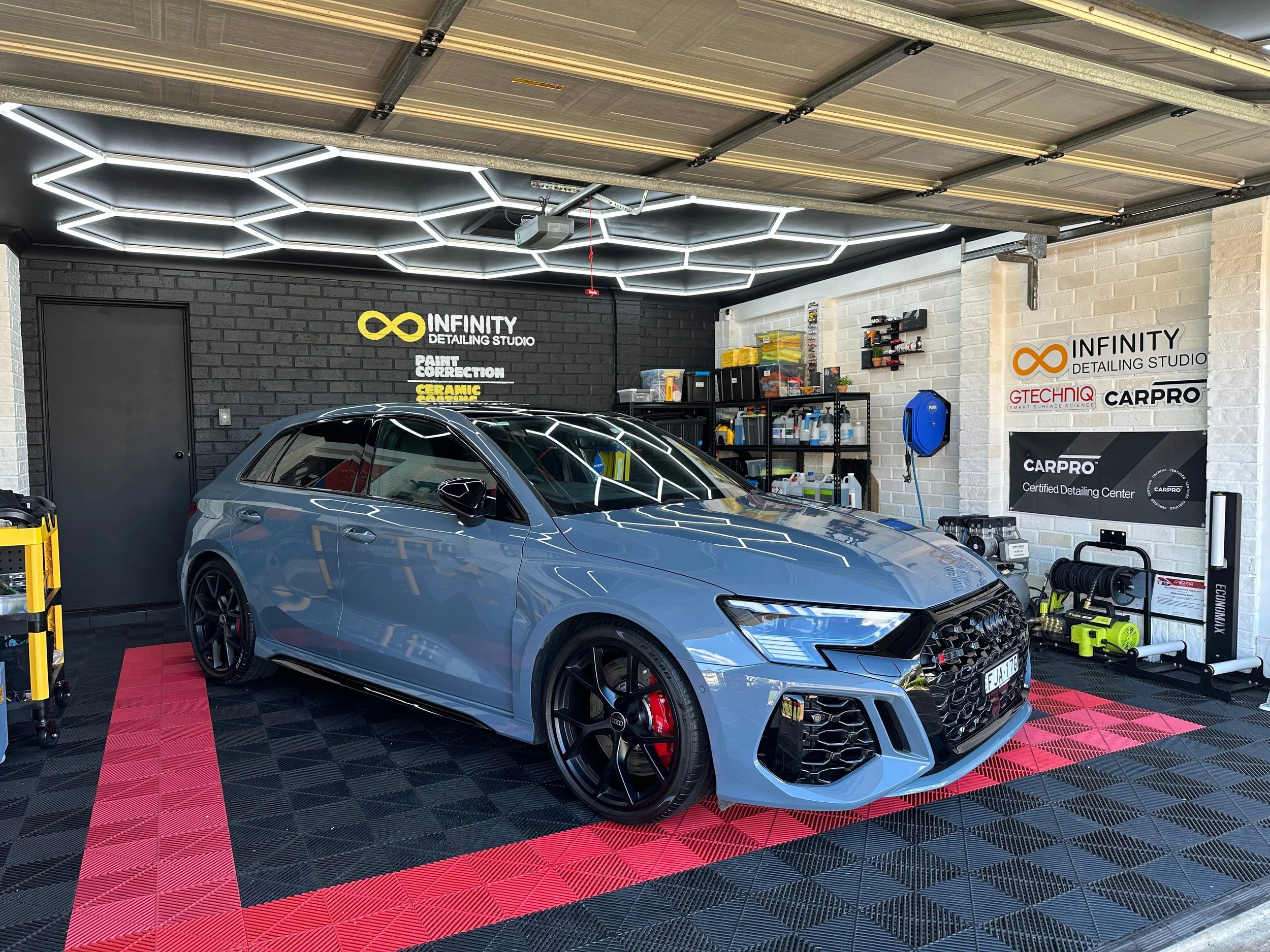 Audi RS3 received an exterior detail, parked on a red and black checkered floor, illuminated ceiling lights, and shelves with detailing supplies. The walls display the logos of Infinity Detailing Studio, Gtechniq, and CarPro.